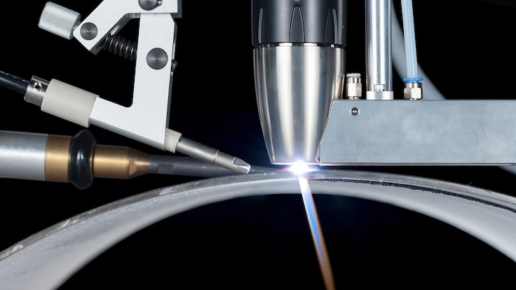 A machine mounted welding torch in action while welding a metal cylinder. The welding torch is producing a bright plasma arc at the metal surface.