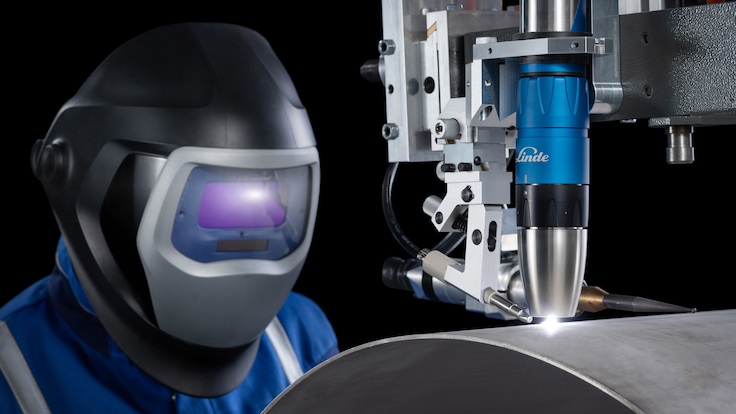 A welder in helmet supervises a machine mounted welding torch in action while welding a metal cylinder. The welding torch is producing a bright plasma arc at the metal surface.
