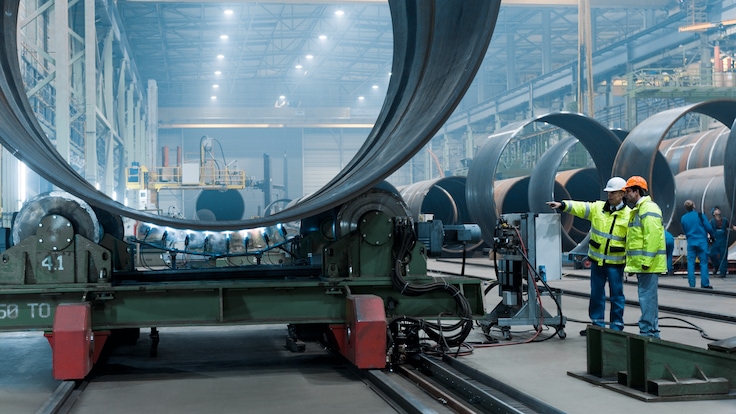 Two workers in personal protective equipment talking and pointing to one of several large cylindrical parts in a wind turbine factory. 