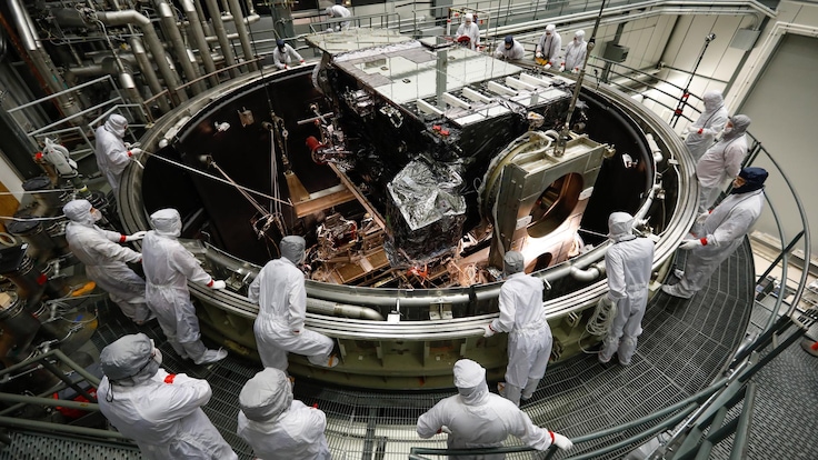 Technicians lowering a satellite into a large round thermal vacuum testing chamber 